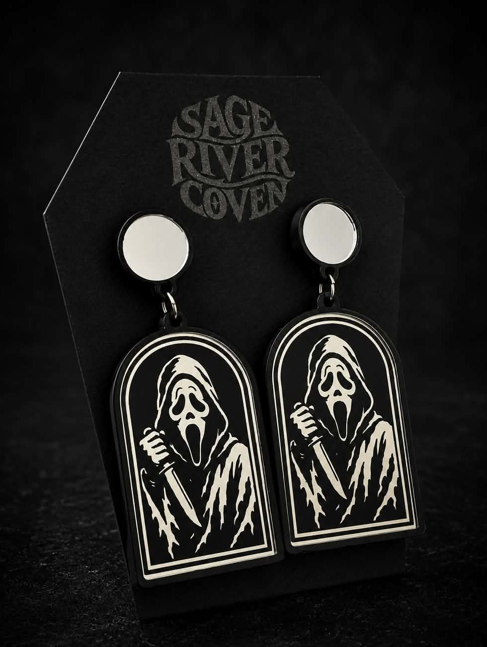 A pair of gothic-style drop earrings displayed on a black coffin-shaped card. The earrings feature a black arched pendant with a white engraved grim reaper design, suspended from round silver studs.