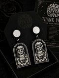A pair of gothic-style drop earrings displayed in a black gift box with a Sage River Coven thank you card in the background. The earrings feature a silver-toned circular top and a black arch-shaped pendant with a white graphic of a skull-faced girl with pigtails holding a rose.