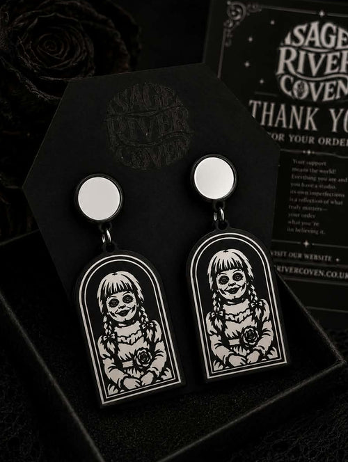 A pair of gothic-style drop earrings displayed in a black gift box with a Sage River Coven thank you card in the background. The earrings feature a silver-toned circular top and a black arch-shaped pendant with a white graphic of a skull-faced girl with pigtails holding a rose.