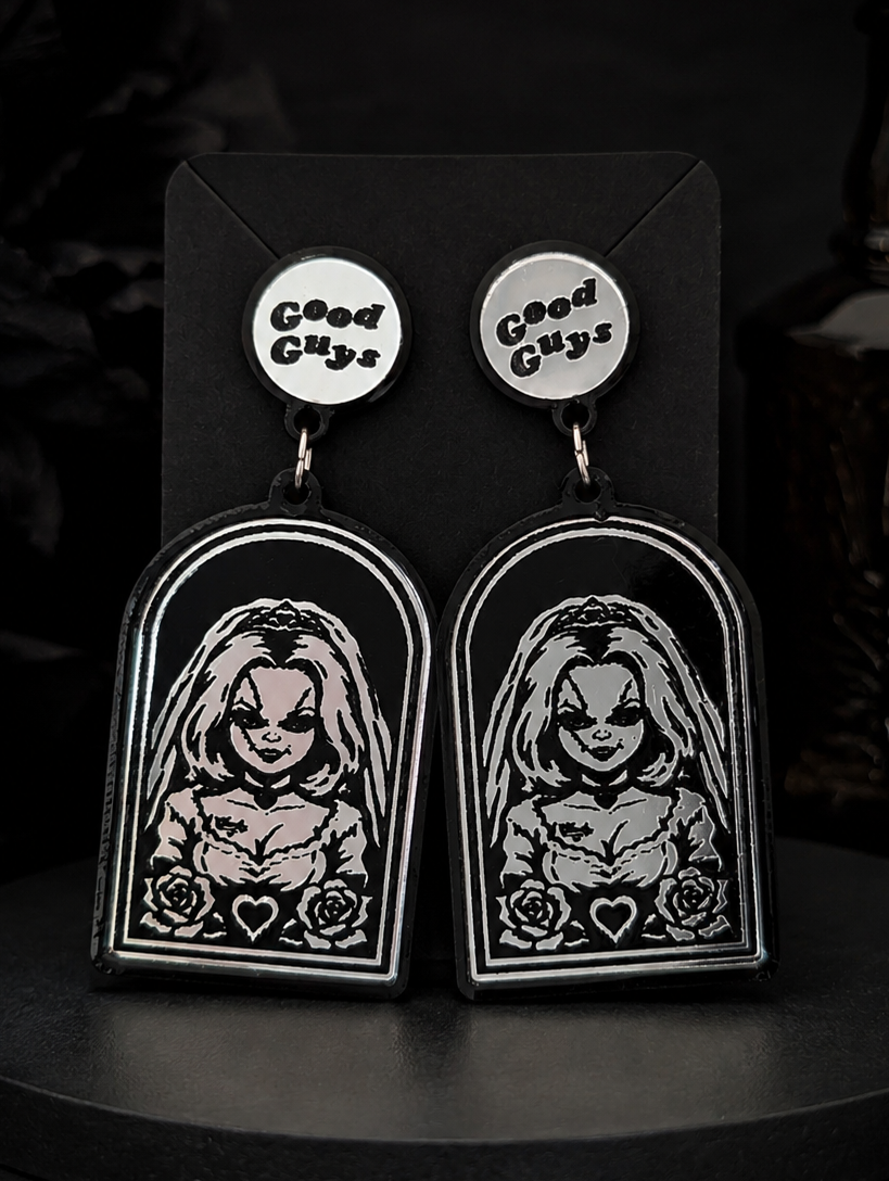 A pair of gothic-style drop earrings displayed on a black card. Each earring has a round silver stud engraved with the words 'Good Guys' and a larger black arch-shaped pendant featuring a creepy illustrated face with long hair, a stitched expression, and small hearts at the bottom.