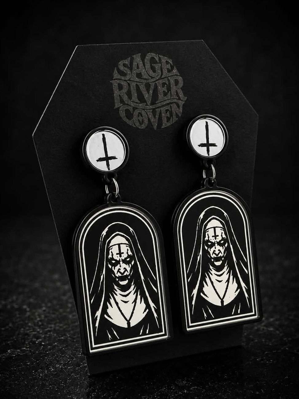 A pair of gothic nun drop earrings displayed on a black coffin-shaped card branded 'Sage River Coven.' The earrings have white oval studs with black symbols and long black arched dangles featuring a high-contrast black-and-white illustration of a hooded figure.